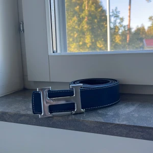 Mörkblått H skärp - Säljer ett mörkblått skärp, i äkta skinn med ikoniskt silverfärgat H-spänne. Bältet har vit kontrastsöm längs kanterna och en stilren, klassisk design som lyfter vilken outfit som helst.