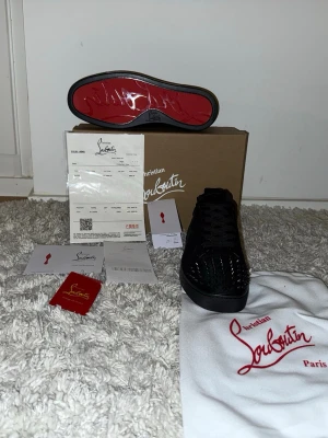 Svarta sneakers från Christian Louboutin - Snygga svarta sneakers från Christian Louboutin med klassisk röd sula och coola nitar framtill. Skorna har snörning och är tillverkade i mocka och skinn, vilket ger en lyxig känsla. Perfekta för dig som vill sticka ut med en exklusiv look.