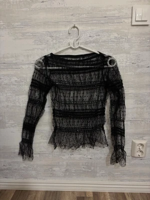 Svart mesh topp med prickigt mönster - Snygg svart mesh topp med lång ärm och prickigt mönster. Toppen har volangdetaljer vid ärmslut och nederkant samt är transparent och figurnära. Perfekt för att skapa en trendig lager-på-lager look.
