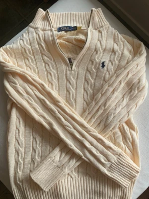 Half zip kabelstickad Ralph Lauren - Kabelstickad beige tröja från Polo Ralph Lauren med halv dragkedja och ribbade muddar. Tröjan har en klassisk passform och den ikoniska blå logotypen broderad på bröstet. Perfekt för lager på lager och snygg till jeans.