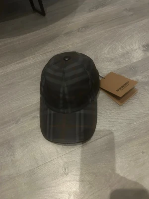 Rutig svart keps från Burberry - Snygg svart keps från Burberry med klassiskt rutigt mönster i mörka toner. Kepsen har böjd skärm och justerbar passform. Perfekt accessoar för att lyfta din outfit med en exklusiv touch. HELT OANVÄND.