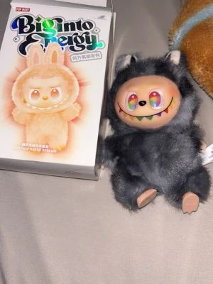 Biginto Energy plush från Pop Mart secret - Säljer en Biginto Energy plush från Pop Mart. Gosedjuret har en fluffig, mörkgrå päls, beige ansikte med regnbågsfärgade ögon och ett stort leende med färgglada tänder. Den har små öron och tassar samt en etikett med The Monsters-loggan. Perfekt för samlare eller som dekoration.