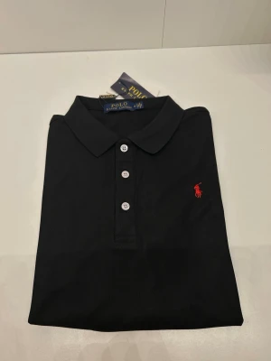 Svart pikétröja från Polo Ralph Lauren - Klassisk svart pikétröja från Polo Ralph Lauren med röd broderad logga på bröstet. Tröjan har krage, tre vita knappar framtill och är tillverkad i bomull. Perfekt för en clean och stilren look. Skriv för annan info
