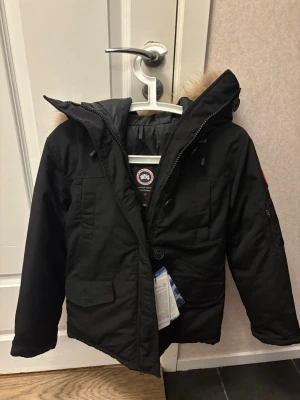 Svart dunjacka från Canada Goose - Snygg svart dunjacka från Canada Goose med luva och beige fuskpälsdetalj. Jackan har dragkedja och knappar framtill, flera fickor och den klassiska loggan på ärmen. Perfekt för kalla vinterdagar och riktigt varm.