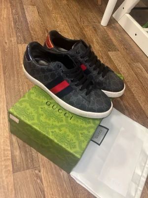 Gucci sneakers med monogram och röd detalj - Snygga svarta Gucci sneakers med klassiskt GG-monogrammönster och röda samt marinblå ränder på sidan. Skorna har vita sulor, svarta skosnören och en kontrasterande röd detalj på hälen. Perfekta för dig som vill ha en lyxig och trendig look.