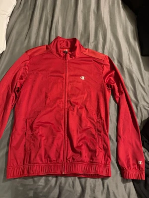 Röd ziptröja från Champion - Röd ziptröja från Champion med liten logga på bröstet och ärmen. Tröjan har hög krage, dragkedja framtill och två fickor. Tillverkad i ett mjukt och lätt material som passar perfekt till vardagslooken.