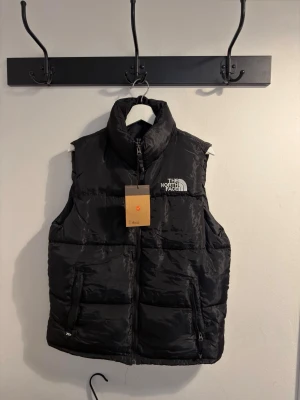 Svart dunväst från The North Face - Stilren svart dunväst från The North Face med hög krage och dragkedja framtill. Västen har två stora fickor med dragkedja och tydlig logga broderad på bröstet. Perfekt för lager-på-lager och håller dig varm under kyliga dagar.