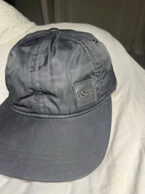 Svart keps från BOSS - Snygg svart keps från BOSS med klassisk böjd skärm och diskret logotyp på sidan. Tillverkad i ett lätt och slitstarkt material som ger en clean och stilren look.