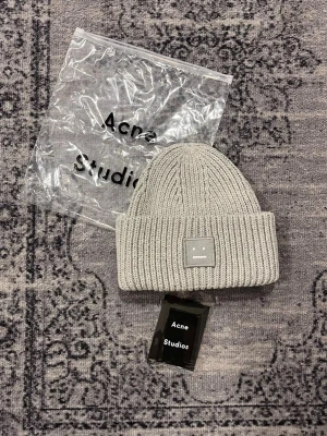 Acne Studios mössa - Säljer en ljusgrå ribbstickad mössa från Acne Studios med uppvikt kant och det klassiska ansiktslogotypen framtill. Mössan är i mjukt och tjockt material, perfekt för kalla dagar. En stilren accessoar som passar till det mesta.