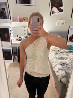 Vit spetsig oneshoulder topp - Snygg vit topp i spets med oneshoulder-design. Toppens material är genombruten spets och den sitter tight mot kroppen. Perfekt för dig som vill sticka ut med en unik och trendig look.
