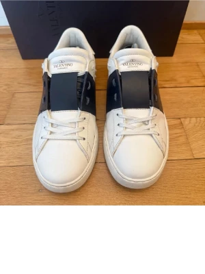 Valentino Open sneakers vit/svart skinn - Snygga Valentino Open sneakers i vitt skinn med bred svart läderdetalj över mitten. Klassisk låg modell med rund tå, vita snören och subtila nitar på hälen. Perfekta för dig som gillar stilrena och exklusiva sneakers.