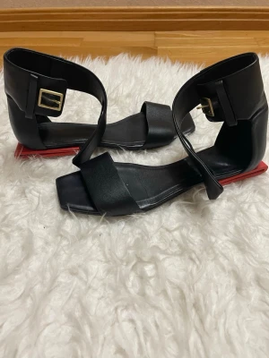 Svarta sandaler med röd detalj - Stilrena svarta sandaler med bred rem över tårna, fyrkantig tå och kraftig ankelrem med guldfärgat spänne. Sulan har en unik röd detalj baktill som sticker ut. Tillverkade i skinnliknande material och har platt sula för en modern look.