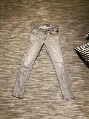 Grå Replay jeans Anbass - Snygga grå jeans från Replay med anbass passform och klassisk femficksdesign. Jeansen är lagade på baksidan men det är knappt synligt när man har på dom. Jeansen har en ljusgrå tvätt, svarta sömmar och tydlig Replay-logga på bakfickan. Perfekta för dig som gillar en smal siluett och stilren look. 