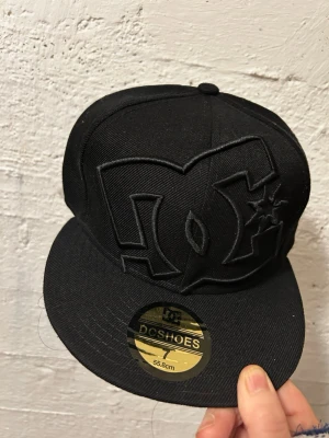Svart DC Shoes keps med broderad logga - Snygg svart keps från DC Shoes med stor broderad logga framtill. Klassisk flat brim-modell med guldfärgad klistermärke på skärmen. Perfekt för dig som gillar streetwear och vill ha en stilren accessoar.
