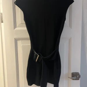 Svart kort klänning med bälte - Stilren svart kort klänning med markerad midja och bälte med metallspänne. Klänningen har en enkel, elegant design med korta ärmar och rak passform. Perfekt för dig som gillar minimalistisk stil. Aldrig använd endast testad 