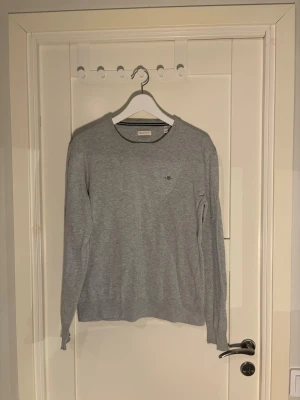 Ljusgrå tröja från GANT - Säljer nu en stilren ljusgrå tröja från GANT med rund halsringning och diskret logga broderad på bröstet. Tröjan har långa ärmar och ribbade muddar vid ärmslut och nederkant. Perfekt för en clean och klassisk look.