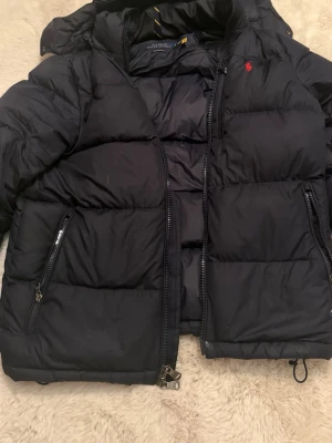 Mörkblå dunjacka från Polo Ralph Lauren - Mörkblå dunjacka från Polo Ralph Lauren med huva och klassisk röd logga på bröstet. Perfekt för kalla dagar och riktigt snygg till streetwear stilen. Jackan har bra skick men ramlade en gång med jackan så det syns lite (se bild5) men det syns knappt irl, annars är den ganska bra skick och har inte använt jackan så mycket 