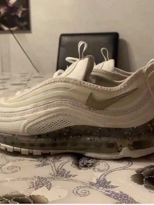 Nike Air Max 97 vita sneakers - Säljer ett par fräscha Nike Air Max 97 sneakers i vitt med ikoniska vågiga linjer och synlig Air-sula. Skorna har snörning och en genomskinlig sula med prickiga detaljer. Perfekta för dig som vill ha en clean och sportig look. Anledning till jag säljer för små och fick present.