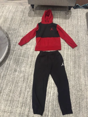 Jordan loungewear set röd/svart - Snyggt loungewear set från Jordan med hoodie och byxor. Hoodien är röd och svart med klassisk Jumpman-logga på bröstet och känguruficka. Byxorna är svarta med vit logga på låret. Perfekt för chill eller sportig stil.