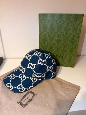 Blå Gucci keps med GG-mönster - Snygg blå keps från Gucci med ikoniskt vitt GG-monogram över hela kepsen. Klassisk böjd skärm och justerbar passform. Tillverkad i slitstarkt tyg och levereras med originalpåse och exklusiv grön presentbox från Gucci.