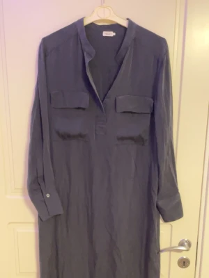 Mörkblå långärmad klänning från Masai - Stilren mörkblå klänning från Masai med lång ärm, v-ringad hals och två bröstfickor. Klänningen har en rak passform och är tillverkad i ett mjukt material som känns skönt mot huden. Perfekt för dig som gillar enkel och avslappnad stil.