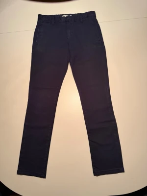 Mörkblå chinos med smal passform - Säljer ett par mörkblå chinos med smal passform och resår i midjan. Byxorna har snedställda fickor fram och två passpoalfickor bak. Perfekta för dig som gillar stilrena och bekväma byxor i bomull. Säljer på grund av att dem är för små. För 12 år.