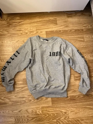 Grå sweatshirt Boohoo med tryck - Grå sweatshirt från Boohoo i storlek M med svart tryck '1984' på bröstet och text på båda ärmarna. Klassisk rund halsringning och ribbade muddar. 