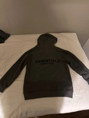 Mörkgrå Essentials Fear of God hoodie - Snygg mörkgrå hoodie från Essentials Fear of God med stor logga på ryggen och mindre logga på bröstet. Klassisk huva och känguruficka framtill. Tillverkad i mjukt material som passar perfekt för en avslappnad streetwear-look.