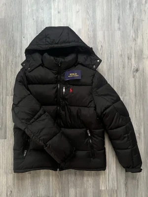 Ralph Lauren jacka  - Snygg svart pufferjacka från Polo Ralph Lauren med huva och dragkedjor framtill. Jackan har det klassiska röda Polo-logot broderat på bröstet och quiltad design. Perfekt för kalla dagar och riktigt stilren look.
