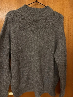 Grå stickad tröja med polokrage - Mysig grå stickad tröja med hög polokrage och lång ärm. Tröjan har en enkel, rak passform och är tillverkad i ett mjukt, tjockt material som passar perfekt för kyliga dagar. Stilren design utan synliga loggor eller mönster.