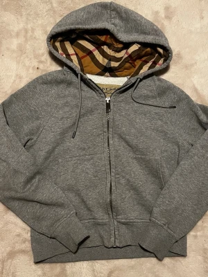 Grå zip hoodie från Burberry dam modell - Säljer en grå zip hoodie från Burberry med klassiskt rutigt foder i huvan. Hoodien har dragsko i huvan, ribbade muddar och fickor framtill. Tillverkad i mjuk bomullsblandning och har en stilren look med ikoniskt Burberry-mönster på insidan av huvan.