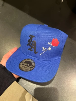 Blå snapback keps med LA och ros - Snygg blå snapback keps från Sport Cap med svart LA-logga och en röd ros broderad på framsidan. Klassisk rak skärm och justerbar baksida. Perfekt för dig som vill sticka ut med en unik och trendig accessoar. Egen designad och helt ny
