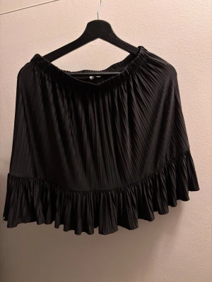 Svart plisserad kort kjol med volang från Zara dam M - Säljer en svart kortkjol med plisserad design och bred volangkant nedtill. Kjolen har resår i midjan för skön passform och är gjord i ett lätt, mjukt material som ger ett snyggt fall. 