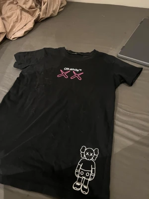 Svart Off-White t-shirt med tryck - Svart t-shirt från Off-White med unikt tryck framtill och stort grafiskt tryck på ryggen. Framsidan har rosa XX och en vit figur, medan baksidan har Off-White-pilar, text och färgdetaljer i lila och rosa. Perfekt för dig som gillar streetwear.