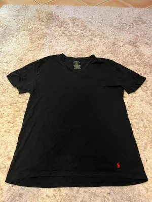 Svart t-shirt från Polo Ralph Lauren - Klassisk svart t-shirt från Polo Ralph Lauren med röd broderad logga nertill. T-shirten har korta ärmar och v-ringad hals. Tillverkad i mjuk bomull som känns skön mot huden. Perfekt basplagg för en clean och stilren look.