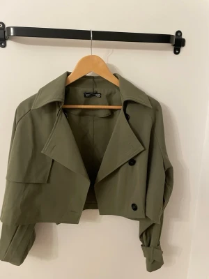 Olivgrön croppad trenchcoat - Snygg croppad trenchcoat i olivgrönt med bred krage och svarta knappar. Jackan har långa ärmar med uppvikta muddar och en klassisk dubbelknäppning framtill. Perfekt för dig som vill ha en trendig och unik jacka till garderoben.