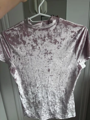 Ljusrosa sammets t-shirt med hög krage - Supersnygg ljusrosa t-shirt i mjuk sammet med korta ärmar och hög krage. Perfekt för dig som vill ha en trendig och unik topp med lite extra lyxig känsla. Passformen är normal och materialet ger ett coolt skimmer.