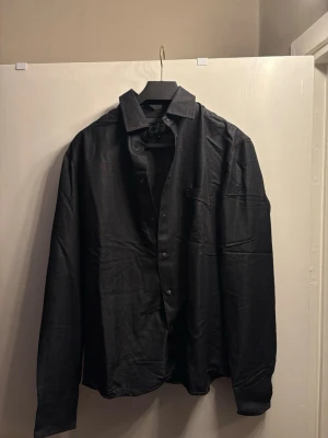 Black Button-Down Shirt with Red Contrast Stitching - Size M - Unique black long-sleeved shirt from the brand d:1. It features a stylish contrast with red stitching and red details on the buttons, giving it a modern and edgy look.  Condition: Excellent condition, sparingly used. Size: Medium (M). Color: Deep black with red thread details. Material: Soft and comfortable high-quality fabric.  Style: Perfect for both semi-formal occasions and a smart-casual daily look. The shirt has a great fit and the red accents really make it stand out from a regular black