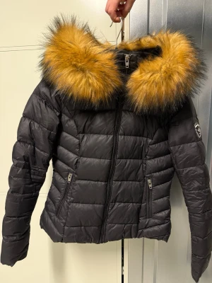 Svart dunjacka Rockandblue med faux fur - Snygg svart dunjacka från Rockandblue med stor fluffig pälskrage i orange och svart. Jackan har quiltad design, dragkedja framtill och två fickor med dragkedja. Märkespatch på ena ärmen ger extra attityd. Perfekt för kalla dagar. Faux fur. Liten i storleken, passar S/M