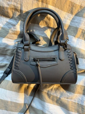 Mörkgrå axelväska med detaljer - Snygg mörkgrå axelväska i syntetmaterial med justerbar axelrem och två rejäla handtag. Väskan har dekorativa sömmar, en liten dragkedjeficka framtill och coola detaljer på sidorna. Väl använd med i gott skick.