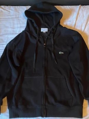 Lacoste Hoodie - Svart hoodie från Lacoste med dragkedja framtill, två fickor och klassisk huva med snören. Ikoniska gröna krokodilloggan broderad på bröstet. Tillverkad i mjukt bomullsmaterial som känns skönt mot huden. Perfekt för en chill och stilren look.                                                         Säljer den för att den inte passar mig längre. Om du är intresserad så kan vi diskutera pris i DM.