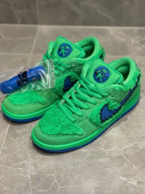 Nike SB Dunk Low Grateful Dead gröna - Unika Nike SB Dunk Low sneakers inspirerade av Grateful Dead, i starkt grön färg med blå detaljer. Skorna har en mix av mocka och fluffigt syntetmaterial, samt ett blått sicksackmönster på swooshen. Tungan pryds av en tecknad björn.