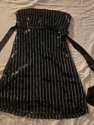Svart kort paljettklänning  - Säljer en supersnygg svart bandeau miniklänning från Miss Harvey med paljetter och ett brett satinband i midjan som knyts till rosett. Perfekt för fest, studentskiva eller liknande!💗