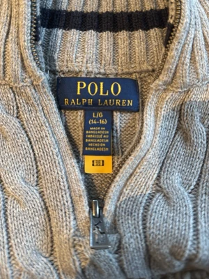 Grå kabelstickad tröja Polo Ralph Lauren - Kabelstickad grå tröja från Polo Ralph Lauren med dragkedja framtill och hög krage. Tröjan har ribbade muddar vid ärmslut och nederkant samt ett klassiskt kabelmönster över hela plagget. Perfekt för dig som gillar stilrena och tidlösa plagg.