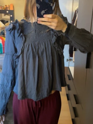 Mörkblå volangblus med spetsdetaljer - Säljer en mörkblå neo noir blus med långa ärmar, volangdetaljer längs axlarna och spetsinslag vid halsringningen och på ärmarna. I väldigt fint skick 🙏🏻