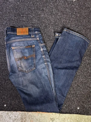 Blå jeans från Nudie Jeans, W29 L32 - Säljer ett par klassiska blå jeans från Nudie Jeans med snygga slitningar och kontrastsömmar. Modellen har fem fickor, orangea detaljer och en ikonisk bakficka med broderad våg. Perfekt passform och riktigt sköna att bära.
