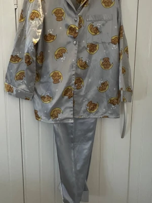 Grå pyjamas med nallemönster från KappAhl - Söt grå pyjamas från KappAhl, Body Zone, med gulligt mönster av bruna nallar som ligger på månar och små vita stjärnor. Pyjamasen har en klassisk krage, bröstficka och knäppning framtill. Materialet är glansigt och består av 83% polyester och 17% bomull.