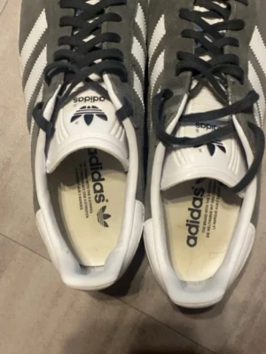 Adidas Gazelle grå/vita sneakers mocka - Säljer ett par klassiska Adidas Gazelle sneakers i grå mocka med vita detaljer och de ikoniska tre ränderna på sidorna. Skorna har svart snörning, vit plös med Adidas-logga och vit sula med mönstrad undersida. Perfekta för dig som gillar retrostil och streetwear.