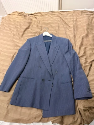 Blå randig kavaj från Oscar Jacobson - Dubbelknäppt blå kavaj från Oscar Jacobson i ull med tunna vita ränder. Klassisk passform med breda slag, fyra knappar på ärmsluten och innerficka. Perfekt för dig som vill ha en stilren och tidlös look. Finns även byxor till för den som önskar!