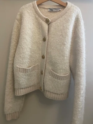 Beige kofta från Zara med fickor - Mysig beige kofta från Zara med fyra guldfärgade knappar framtill. Två stora fickor och ribbade muddar vid ärmslut och nederkant. Perfekt nu när det är lite mer kallt ute men ändå med stil.